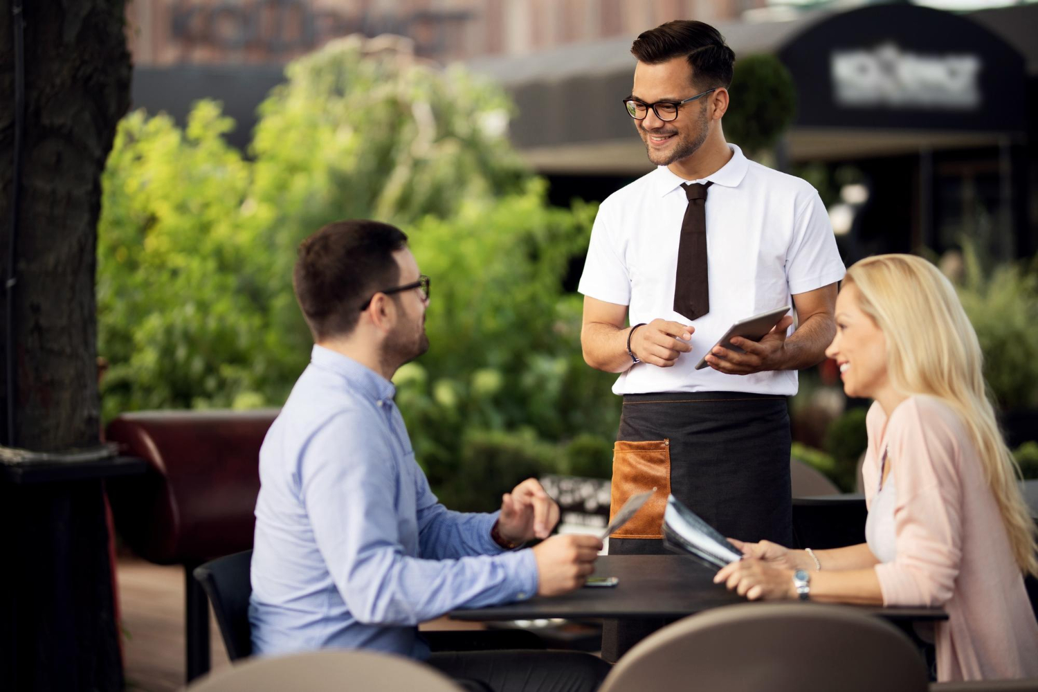 Serveur utilisant un pad de commande en terrasse Serveur souriant prenant une commande avec un pad numérique auprès de deux clients en terrasse dans un restaurant moderne.